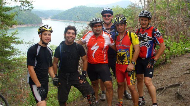 A group of six men in cycling gear, posing together with mountain bikes beside a scenic lake surrounded by trees and hills in the background. The atmosphere is casual and friendly, showcasing a moment from a biking adventure. Tsali Right Loop mountain bike trail.
