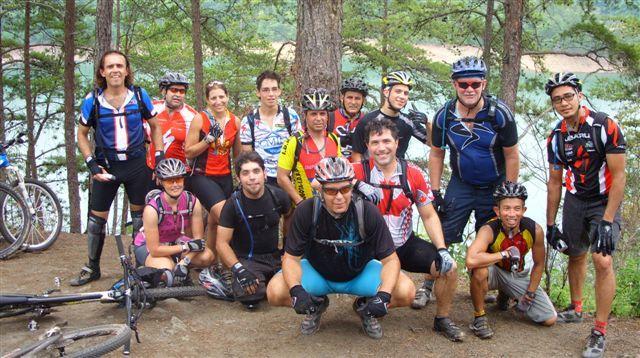 A group of 16 mountain bikers posing together in a forested area near a lake. They are wearing colorful cycling jerseys and helmets, and some bikes are parked nearby. The group is smiling and making gestures, enjoying a break during a biking adventure. Tsali Right Loop mountain bike trail.
