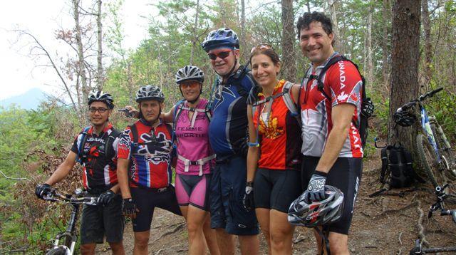 A group of six mountain bikers pose together for a photo on a forest trail. They are wearing colorful cycling jerseys and helmets, surrounded by trees and nature. Some bicycles are visible in the background. The group appears cheerful and engaged in outdoor activity. Tsali Right Loop mountain bike trail.
