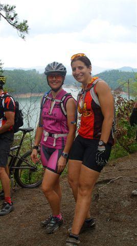 Two women are standing on a trail beside a lake, smiling at the camera. They are both wearing cycling gear: one in a pink top with the words "Team" and the other in a red and black outfit. In the background, there are trees and a glimpse of the lake under a cloudy sky. Other cyclists are visible nearby. Tsali Right Loop mountain bike trail.