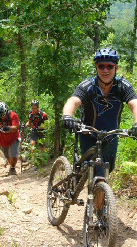 Three mountain bikers ascend a rocky, wooded trail. The focus is on a smiling cyclist in the foreground, wearing a black helmet and sunglasses, as he pedals up the path. Two other cyclists follow behind, one wearing a red shirt and the other in a black and red outfit. Lush green foliage surrounds them, indicating a vibrant outdoor setting. Tsali Right Loop mountain bike trail.