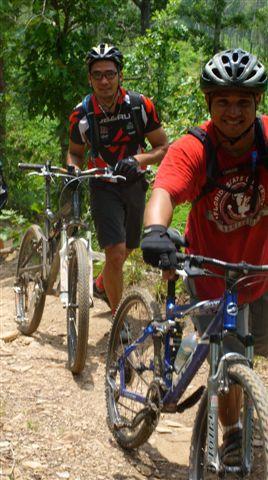 Two cyclists are climbing a dirt trail surrounded by lush greenery. One cyclist is wearing a red shirt and smiling at the camera, while the other, dressed in a black and red outfit, follows closely behind. Both are riding mountain bikes. Tsali Right Loop mountain bike trail.