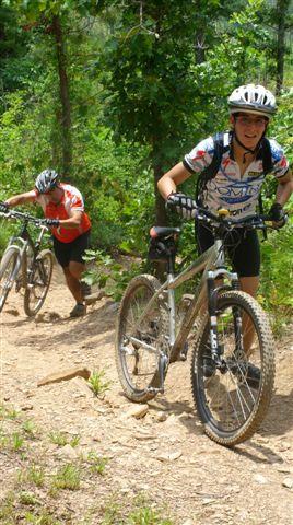 Two mountain bikers are navigating a dirt trail in a lush, green forest. One biker is pushing their bike uphill, while the other is riding, both wearing helmets and cycling attire. The scene captures the challenging terrain and the outdoor activity. Tsali Right Loop mountain bike trail.