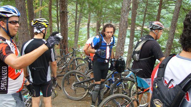 A group of mountain bikers gathered on a forest trail, surrounded by trees. Several cyclists are seen adjusting their gear and bikes, with some bikes parked nearby. The setting suggests an outdoor activity, likely after a ride or at a trailhead, with a glimpse of a body of water in the background. Tsali Right Loop mountain bike trail.