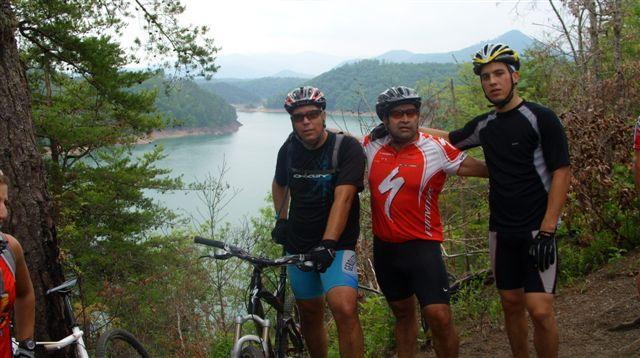Three male mountain bikers pose for a photo on a scenic overlook near a tranquil lake surrounded by green hills. They are wearing cycling gear and helmets, with bicycles parked nearby. The background features a calm water body and distant mountains under a cloudy sky. Tsali Right Loop mountain bike trail.