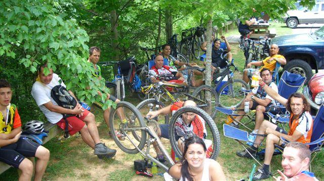 A group of cyclists resting in a shaded area, surrounded by trees and parked bicycles. Some people are sitting on the ground, while others are in camping chairs. They are dressed in colorful cycling jerseys and appear to be enjoying a break during a biking event or outing. A vehicle is visible in the background. Tsali Right Loop mountain bike trail.