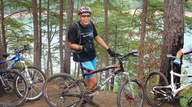 A person wearing a black shirt and blue shorts stands next to a mountain bike in a forested area. In the background, several trees and a body of water are visible. The cyclist is smiling and wearing a helmet and gloves, suggesting an outdoor biking activity. Other bikes can be seen nearby. Tsali Right Loop mountain bike trail.