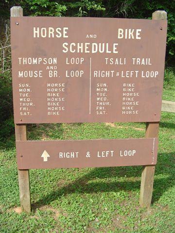 A wooden sign displaying a schedule for horse and bike activities on trails, including Thompson Loop, Mouse Branch Loop, Tsali Trail, and Right & Left Loop. The sign lists days of the week with designated activities for horses and bikes. An arrow at the bottom indicates directions for the Right & Left Loop. Tsali Thompson Loop mountain bike trail.