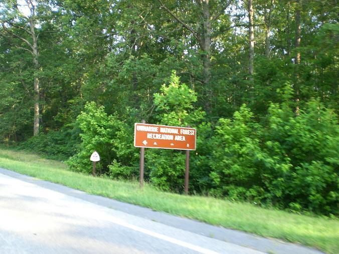 A roadside sign indicating the entrance to the Ouachita National Forest Recreational Area, surrounded by lush green foliage and trees. Uwharrie NF: Wood Run, Supertree And Keyauwee mountain bike trail.