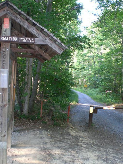 A wooden information kiosk at the entrance of a trailhead surrounded by lush green trees, with signs pointing to "Information" and "Mountain Biking." A gravel path leads into the woods, and a barrier gate is visible in the foreground. Uwharrie NF: Wood Run, Supertree And Keyauwee mountain bike trail.