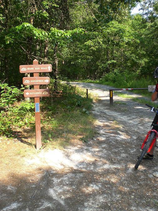A dirt path leading into a wooded area, marked by wooden trail signs pointing in different directions: "Wood Run Road," "Rope Bridge," and "Keyavale." A person with a bicycle is visible on the right, near a barrier at the entrance to the trail. Lush green trees surround the scene, indicating a natural outdoor environment. Uwharrie NF: Wood Run, Supertree And Keyauwee mountain bike trail.