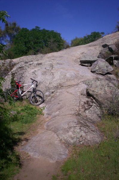 A mountain bike parked against a large rocky slope, surrounded by greenery and blue sky. The terrain is rugged with a mix of flat rock and boulders, indicating a natural outdoor setting ideal for biking or hiking. Granite Bay Trail mountain bike trail.