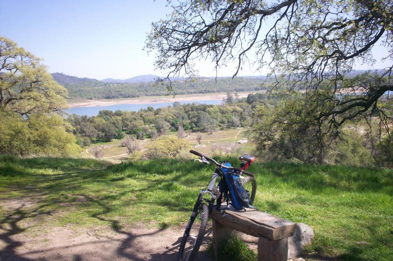 A scenic view overlooking a reservoir surrounded by lush greenery and hills, with a mountain bike resting on a wooden bench in the foreground. Sunlight filters through the trees, creating a tranquil atmosphere perfect for outdoor activities. Granite Bay Trail mountain bike trail.