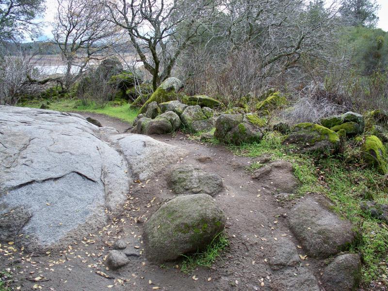 A rugged pathway winding through a serene landscape featuring large, moss-covered boulders, patches of green grass, and leafless trees in the background. The scene is set against a cloudy sky, suggesting a tranquil, natural environment. Granite Bay Trail mountain bike trail.