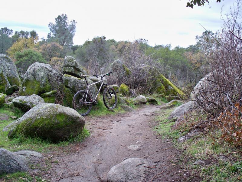 A mountain bike parked beside a dirt path, surrounded by large rocks and sparse vegetation, with a cloudy sky in the background. Granite Bay Trail mountain bike trail.