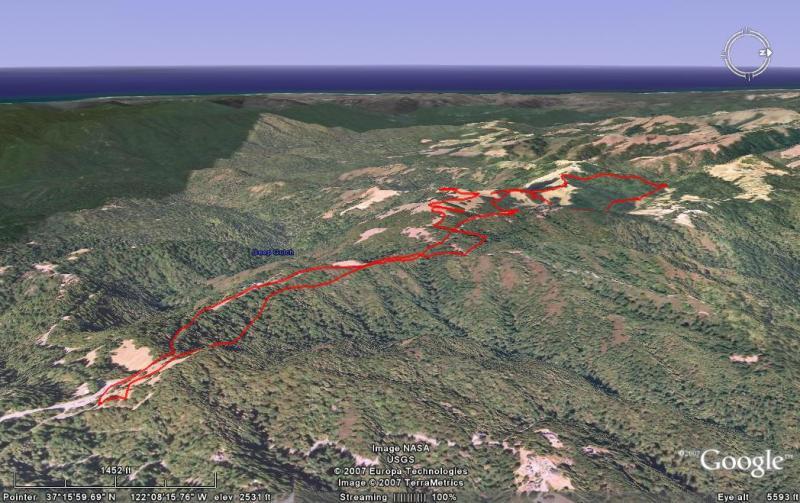 A 3D satellite view of a mountainous landscape, featuring a winding red route that traces a hiking or trail path through dense green forests. The ocean is visible in the distance, and the image includes geographical labels and coordinates. Saratoga Gap mountain bike trail.