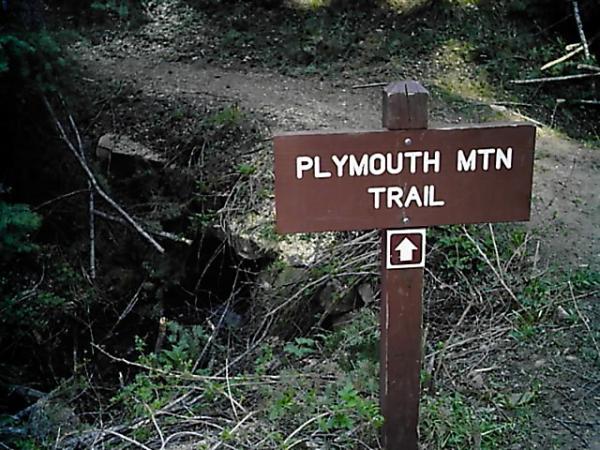 A wooden sign for the Plymouth Mountain Trail, pointing in the direction of the trail, surrounded by greenery and a dirt path in the background. Deer Creek Canyon mountain bike trail.