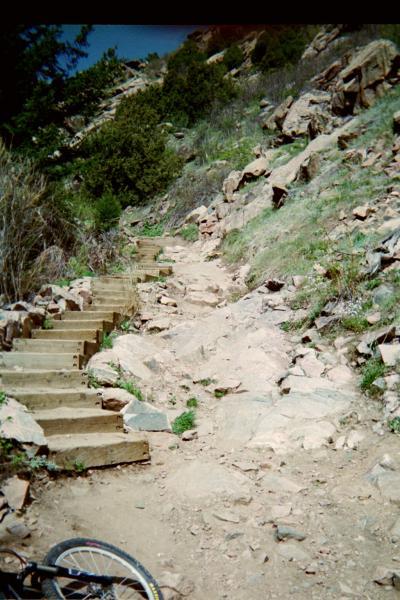A rocky hiking path with wooden steps leading up a hill, surrounded by greenery and boulders. A bike is partially visible in the foreground. Deer Creek Canyon mountain bike trail.