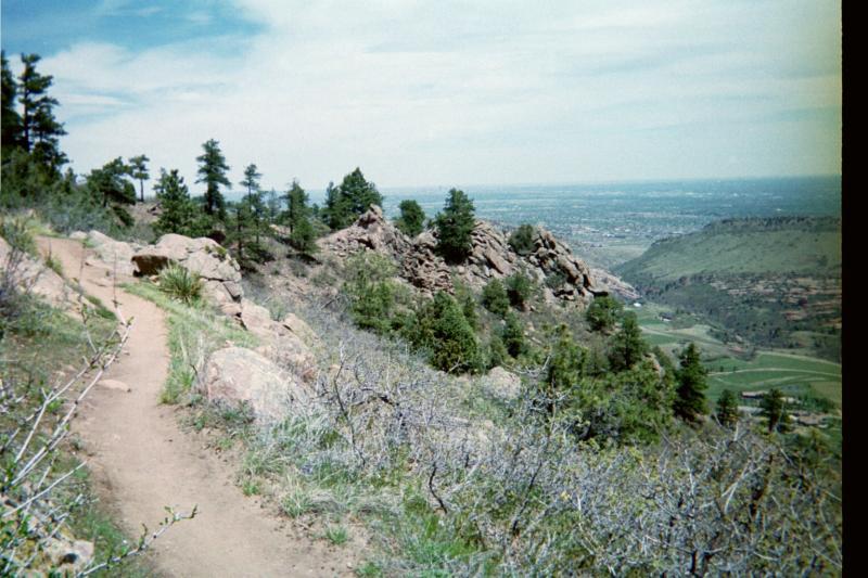 A rocky trail winds along a hillside, surrounded by lush greenery and pine trees. In the distance, a panoramic view reveals valleys and a sprawling landscape under a partly cloudy sky. Deer Creek Canyon mountain bike trail.