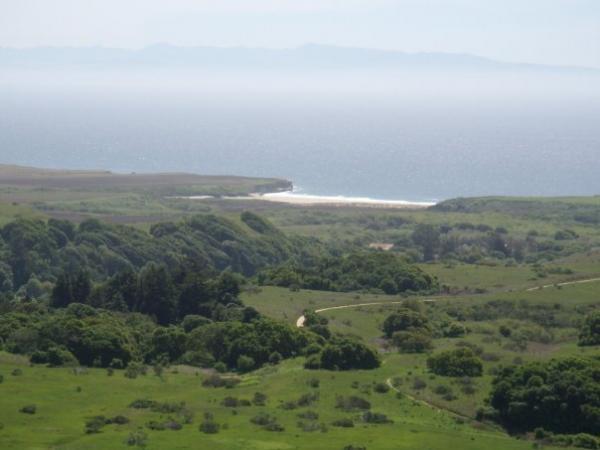 A scenic view overlooking a coastal landscape, featuring rolling green hills, lush vegetation, and a shoreline meeting the ocean in the distance. The calm sea reflects the sunlight, with a faint outline of mountains visible on the horizon under a clear sky. Wilder Ranch State Park mountain bike trail.