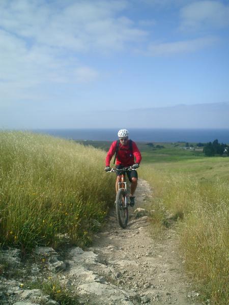 A mountain biker in a red jacket rides along a rocky trail, surrounded by tall grasses and a scenic view of the ocean in the background under a partly cloudy sky. Wilder Ranch State Park mountain bike trail.