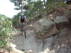 A mountain biker riding on a narrow, sandy trail surrounded by rocks and greenery, with a slight jump over a dip in the path. Palmer Park mountain bike trail.