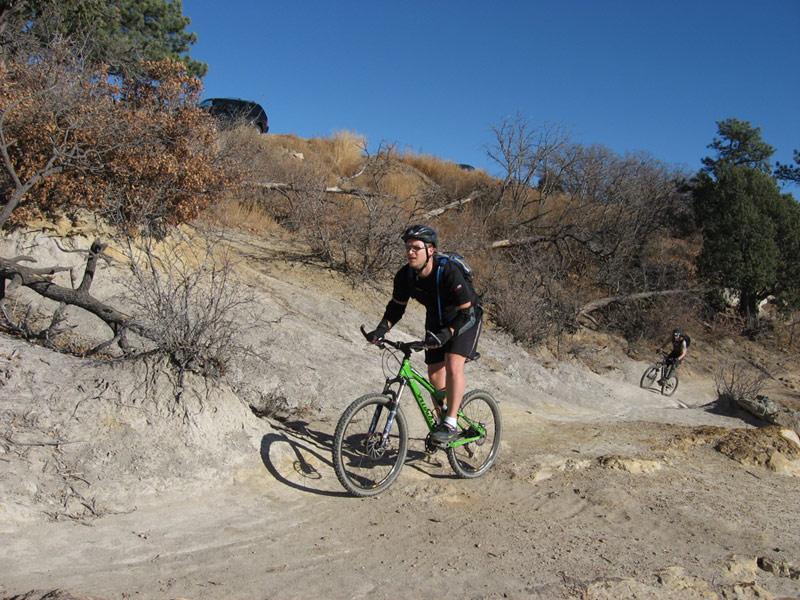 A mountain biker riding a green bicycle on a rocky trail, with a second biker visible in the background. The setting features dry, hilly terrain with sparse vegetation and blue skies. Palmer Park mountain bike trail.
