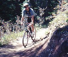 A person riding a mountain bike along a dirt trail surrounded by greenery and trees. The rider is wearing a helmet and casual athletic clothing, demonstrating an active outdoor lifestyle. Waterton Canyon mountain bike trail.