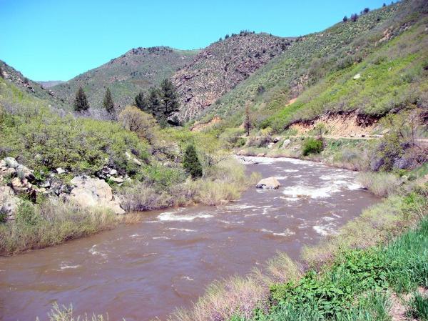 A winding river flows through a lush green valley surrounded by mountains. The landscape features a mix of rocky outcrops and dense vegetation, with the river's water appearing brown and slightly turbulent, likely from recent rainfall. Bright blue skies overhead complement the vibrant greenery. Waterton Canyon mountain bike trail.