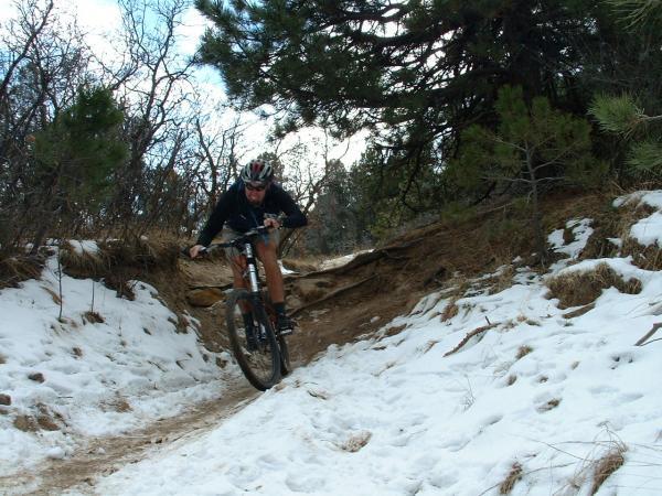 A mountain biker navigating a snowy trail, performing a jump over a section of the path surrounded by trees and shrubs. Palmer Park mountain bike trail.