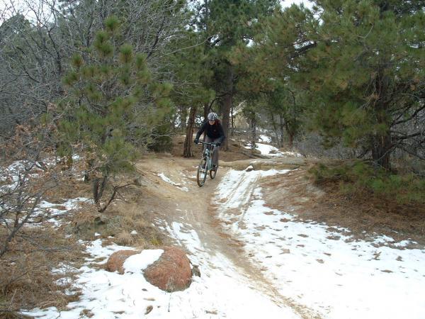 A mountain biker navigating a snowy trail surrounded by pine trees, with rocky terrain and patches of bare ground visible. The cyclist is in motion, wearing a helmet and dark clothing. Palmer Park mountain bike trail.