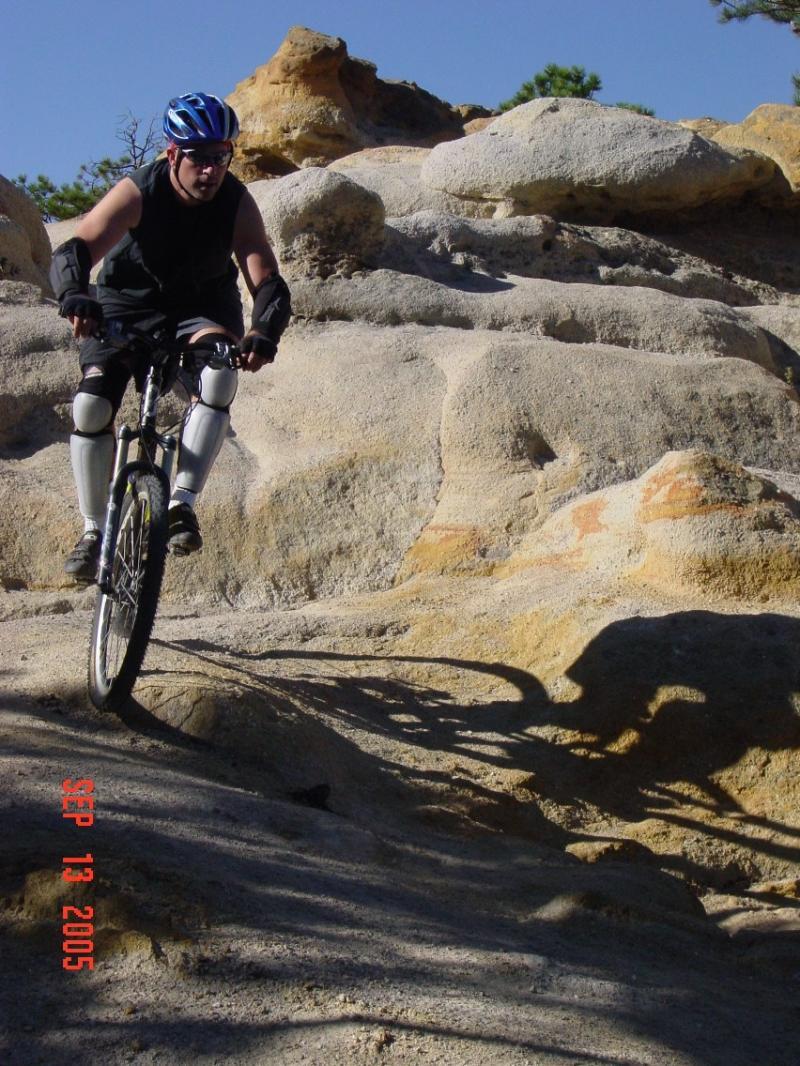 A person in protective gear riding a mountain bike on rocky terrain, with a clear blue sky in the background. The rider is focused as they navigate the uneven surface, casting a shadow on the ground. Palmer Park mountain bike trail.