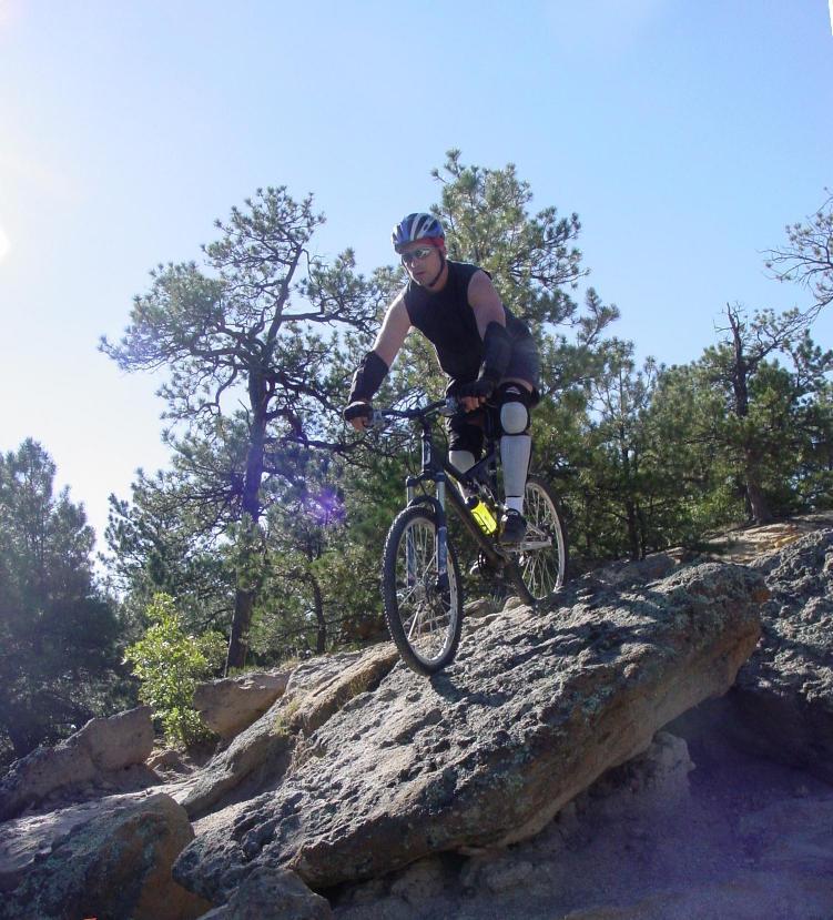A cyclist skillfully navigating a rocky trail on a mountain bike, wearing a helmet and protective gear, surrounded by trees and clear blue skies. Palmer Park mountain bike trail.