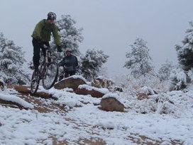A mountain biker in a green jacket performs a jump on a snowy trail, with snow-covered rocks and trees in the background. Another biker can be seen in the distance. Palmer Park mountain bike trail.