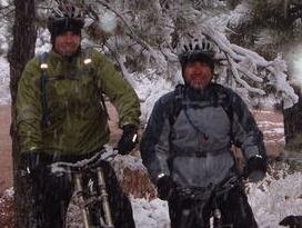 Two men wearing bicycle helmets and winter clothing stand next to their mountain bikes in a snowy forested area. Snow is falling, creating a wintry atmosphere around them. Palmer Park mountain bike trail.