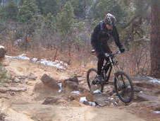 A person wearing a helmet and biking gear rides a mountain bike on a rocky trail in a forested area, with patches of snow on the ground and trees in the background. Palmer Park mountain bike trail.