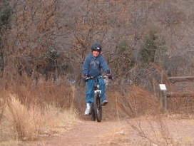 A person in a blue jacket and helmet riding a bicycle on a dirt trail surrounded by dry grass and sparse trees. Palmer Park mountain bike trail.