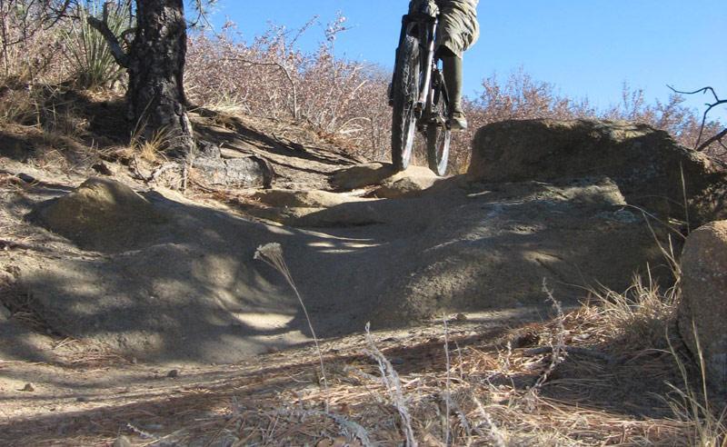 A mountain biker navigating a rocky trail, with the bike's back wheel elevated above the ground, surrounded by dry vegetation and blue sky. Palmer Park mountain bike trail.