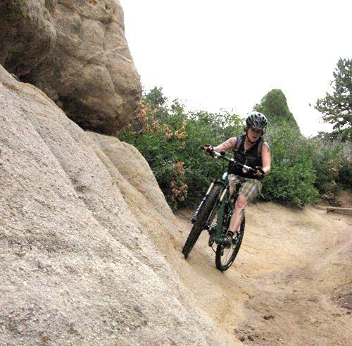 A mountain biker navigating a rocky trail surrounded by greenery, wearing a helmet and casual clothing. The terrain features steep, uneven surfaces and natural rock formations. Palmer Park mountain bike trail.