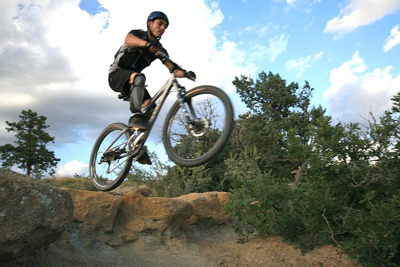 A mountain biker in protective gear jumps over a rock formation on a trail, with trees and a partly cloudy sky in the background. Palmer Park mountain bike trail.