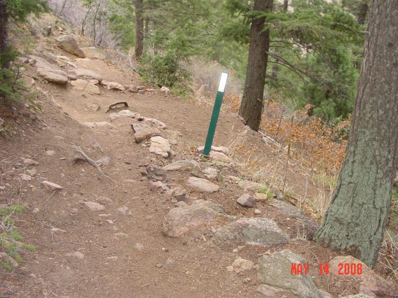 A narrow dirt trail winding through a forest, featuring rocky sections and lush green trees. A vertical green post with a reflective top stands along the trail, indicating the path. The scene is set in a natural environment, with bare branches and a hint of autumn foliage in the background. The date in the corner reads May 14, 2006. Palmer Trail / Section 16 mountain bike trail.