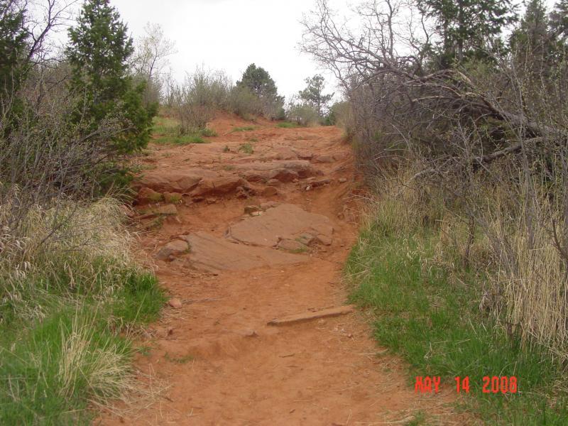 A rugged dirt trail surrounded by sparse vegetation and rocky terrain, leading upward under a cloudy sky. The path features exposed red earth and scattered rocks, with patches of grass and small bushes along the sides. Palmer Trail / Section 16 mountain bike trail.