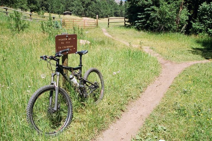 A mountain bike is leaned against a trail sign, surrounded by tall green grass and wildflowers. The sign indicates a path leading to Colorado, and in the background, there is a forested area and an open field with a fence. Two visible trails diverge from the sign, inviting exploration into the natural landscape. Colorado Trail: Green Mountain mountain bike trail.
