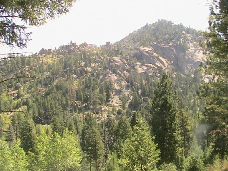 A scenic view of a mountainous landscape, featuring rugged rock formations and dense evergreen forests under a bright, sunny sky. The slopes are covered with a mix of trees, showcasing vibrant greens. Colorado Trail: Green Mountain mountain bike trail.
