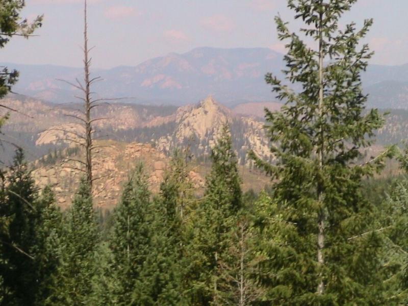 A scenic mountain landscape featuring dense evergreen trees in the foreground, rocky formations in the midground, and distant mountains under a clear sky. Colorado Trail: Green Mountain mountain bike trail.