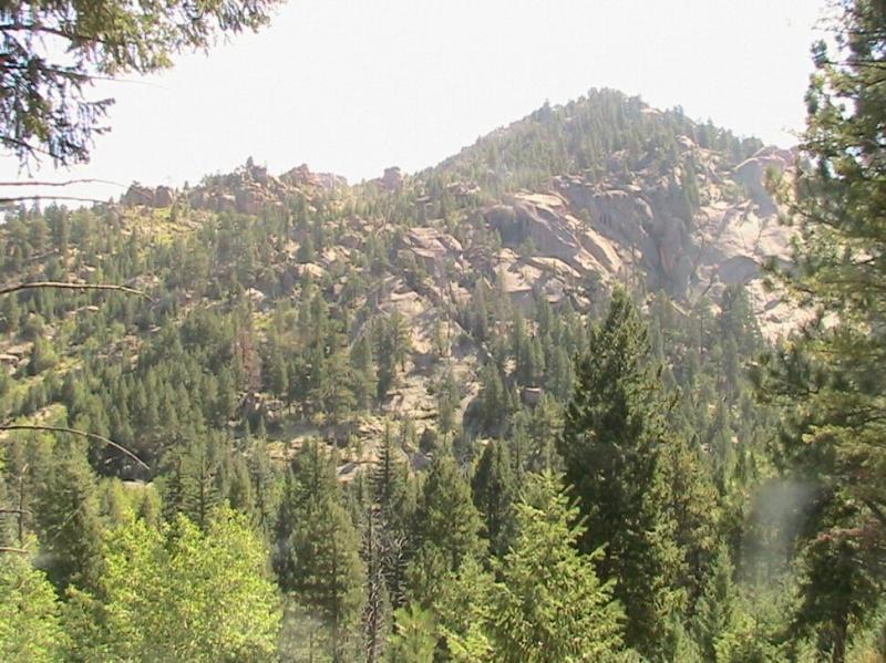 A scenic view of rocky mountains surrounded by dense greenery, featuring a variety of trees under bright sunlight. The landscape showcases the natural beauty of the wilderness, with steep rock formations and lush foliage in the foreground. Colorado Trail: Green Mountain mountain bike trail.