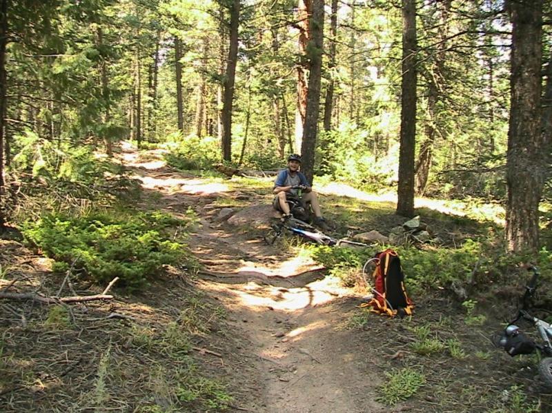 A person sitting on a rocky path in a forested area, surrounded by trees and greenery. A bicycle is propped up nearby, and a backpack lies on the ground. The scene is sunny and serene, suggesting a peaceful moment during a biking or hiking adventure. Colorado Trail: Green Mountain mountain bike trail.