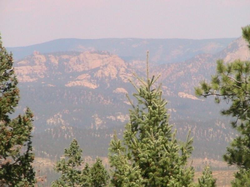 A scenic view of a mountainous landscape, featuring towering rock formations in the background, partially shrouded by a haze. In the foreground, there are several green pine trees, adding depth to the natural setting. The overall atmosphere conveys a sense of tranquility and natural beauty. Colorado Trail: Green Mountain mountain bike trail.