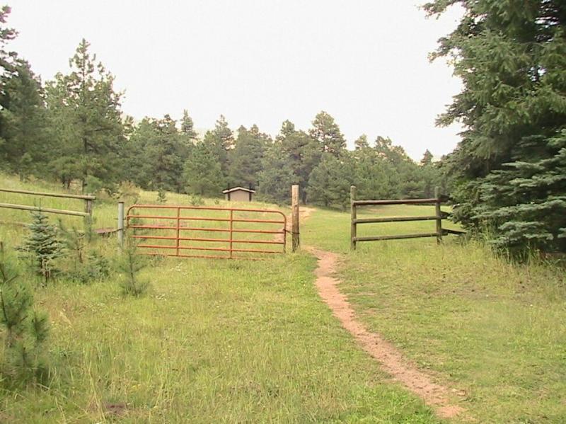 A dirt path leading through an open gate into a grassy area, surrounded by trees. A red metal gate is on the left, and a wooden fence is on the right, with a small structure visible in the background. The scene is framed by greenery, creating a serene outdoor setting. Colorado Trail: Green Mountain mountain bike trail.