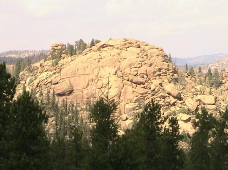 A large, rocky outcrop surrounded by evergreen trees, set against a backdrop of distant mountains under a partly cloudy sky. The rocky surface is composed of light-colored boulders, showcasing natural geological formations. Colorado Trail: Green Mountain mountain bike trail.